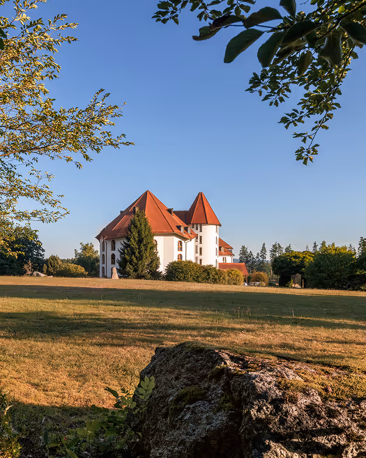 Weißes Schloss mit roten Dächern in einer weiten Wiese, umgeben von Bäumen unter klarem blauem Himmel.