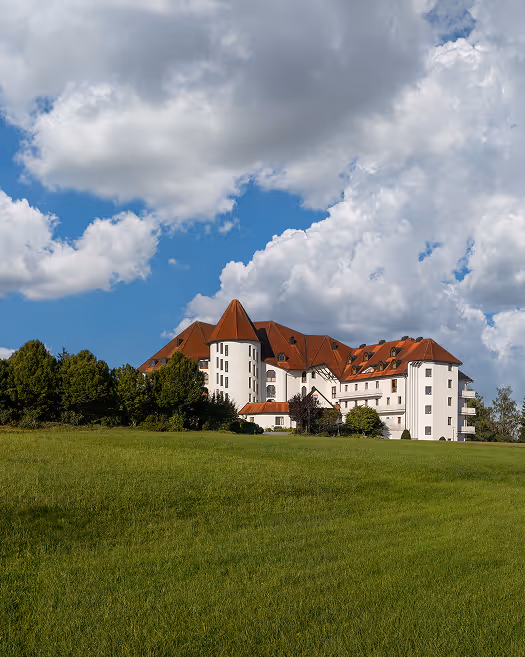 Schloss auf einem Hügel mit weißen Wänden, roten Dächern, umgeben von Bäumen und einer grünen Wiese unter einem bewölkten blauen Himmel.
