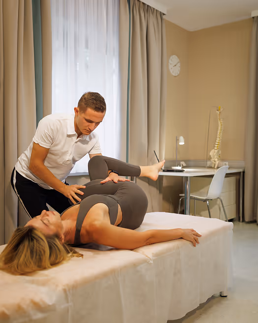 Physiotherapeut in weißem Polo dehnt Patientin in grauer Trainingskleidung auf Behandlungsliege.