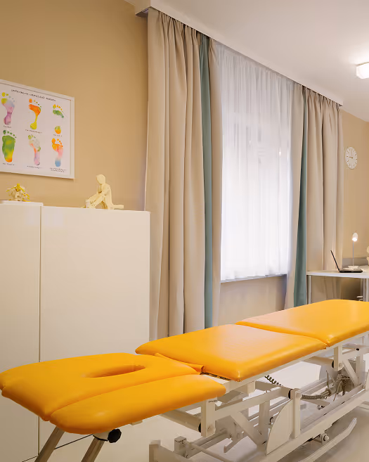 Behandlungsraum mit gelber Massageliege, weißen Schränken und einem Fußreflexzonen-Poster an der Wand.