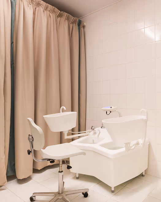 Weiße, medizinische Fußpflegeausrüstung mit einem rollbaren Stuhl vor beigefarbenen Vorhängen und weißen Kacheln.