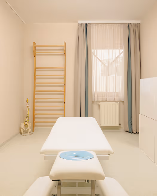 Behandlungsraum mit weißer Liege, Fenster mit Vorhängen und Holzleiter an der Wand.