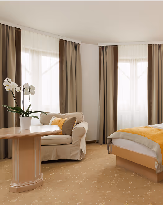 Helles Hotelzimmer mit beigem Sessel, Holztisch mit weißer Orchidee, großen Fenstern mit braunen Vorhängen und Bett mit gelber Decke.