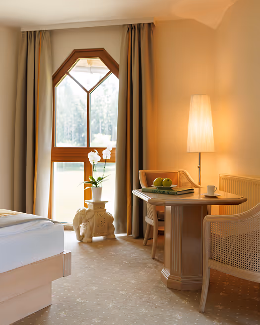 Helles Hotelzimmer mit Holztisch, zwei Stühlen, einer Lampe und einem Fenster mit Vorhängen.