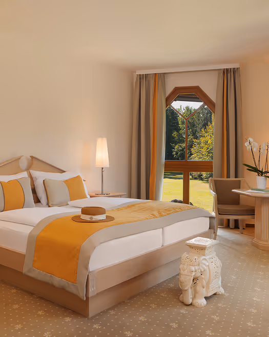 Hell eingerichtetes Hotelzimmer mit Doppelbett, gelb-grauer Bettdecke, Nachttischlampe, Fenster mit Vorhängen und Blick auf grüne Bäume.