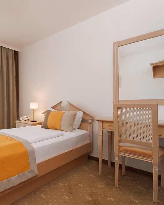 Ein schlichtes Hotelzimmer mit Einzelbett, gelben und grauen Kissen, Schreibtisch mit Stuhl und Nachttisch mit Lampe.