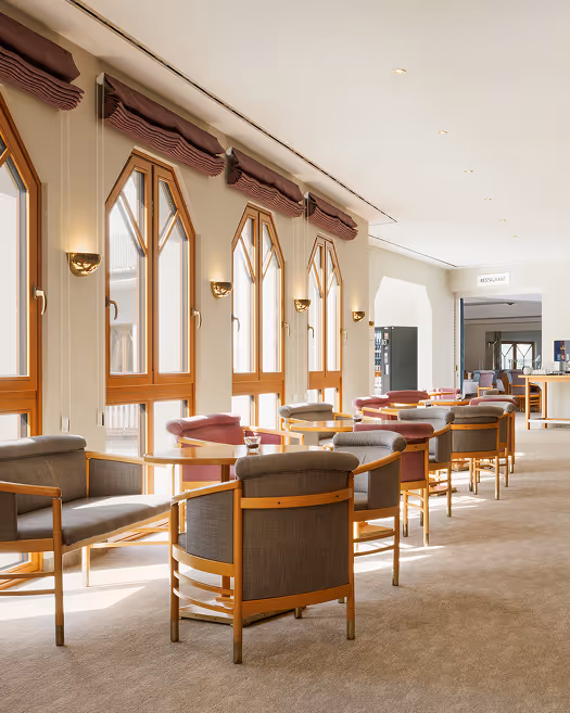 Hell beleuchteter Raum mit mehreren Sitzgruppen aus grauen und roten Sesseln und Holztischen neben großen Fensterfronten.