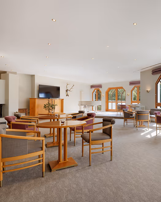 Helles, geräumiges Wohnzimmer mit mehreren Tischen und Stühlen aus Holz, großen Fenstern und einem Fernseher an der Wand.