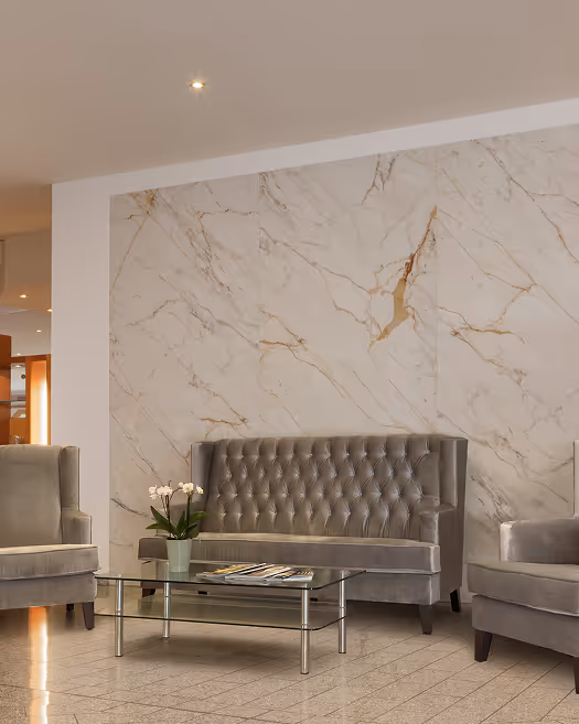 Modernes Wohnzimmer mit grauem Samtsofa, zwei Sesseln, Glas-Couchtisch und Topf mit weißen Orchideen vor einer Wand aus Marmor.