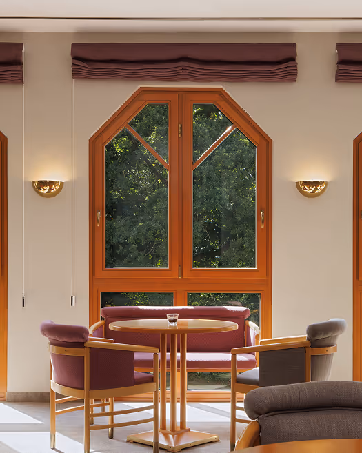 Gemütlicher Sitzbereich mit runder Holztisch, gepolsterten Stühlen und Sofa vor einem großen Fenster mit Blick auf grüne Bäume.