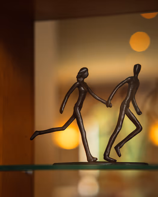 Skulptur zweier abstrakter Figuren, die sich an den Händen halten, wobei eine Person dem anderen folgt.