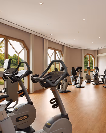 Innenraum eines Fitnessstudios mit mehreren stationären Fahrrädern vor großen Fenstern und hellen Holzböden.