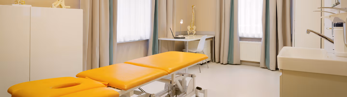 Behandlungszimmer mit orangefarbenem Untersuchungstisch, Schreibtisch mit Wirbelsäulenmodell und Stuhl, sowie Waschbecken.