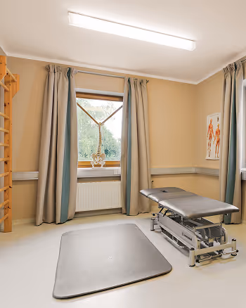 Behandlungsraum mit einer höhenverstellbaren Massagebank, einer Gymnastikmatte auf dem Boden und anatomischem Poster an der Wand.