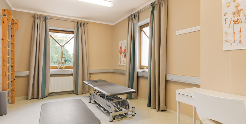 Behandlungszimmer mit Untersuchungsliege, zwei Fenstern mit grauen Vorhängen, anatomischen Postern an der Wand und einem weißen Schreibtisch mit Stuhl.