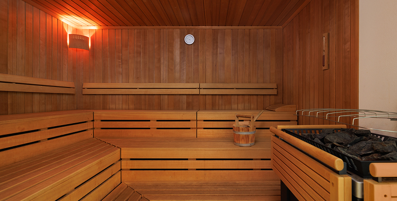 Holzsauna mit mehrstufigen Sitzbänken, einem Eimer mit Kelle und Ofen mit aufgeheizten Steinen.
