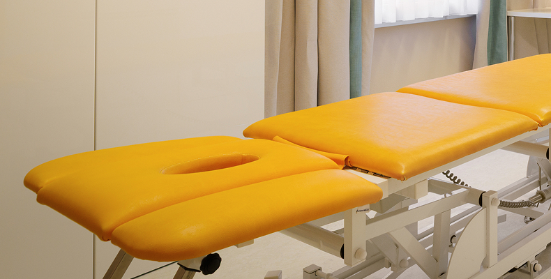 Gelbes Behandlungsliegenbett in einem hellen Therapieraum mit Vorhängen und Fenster.