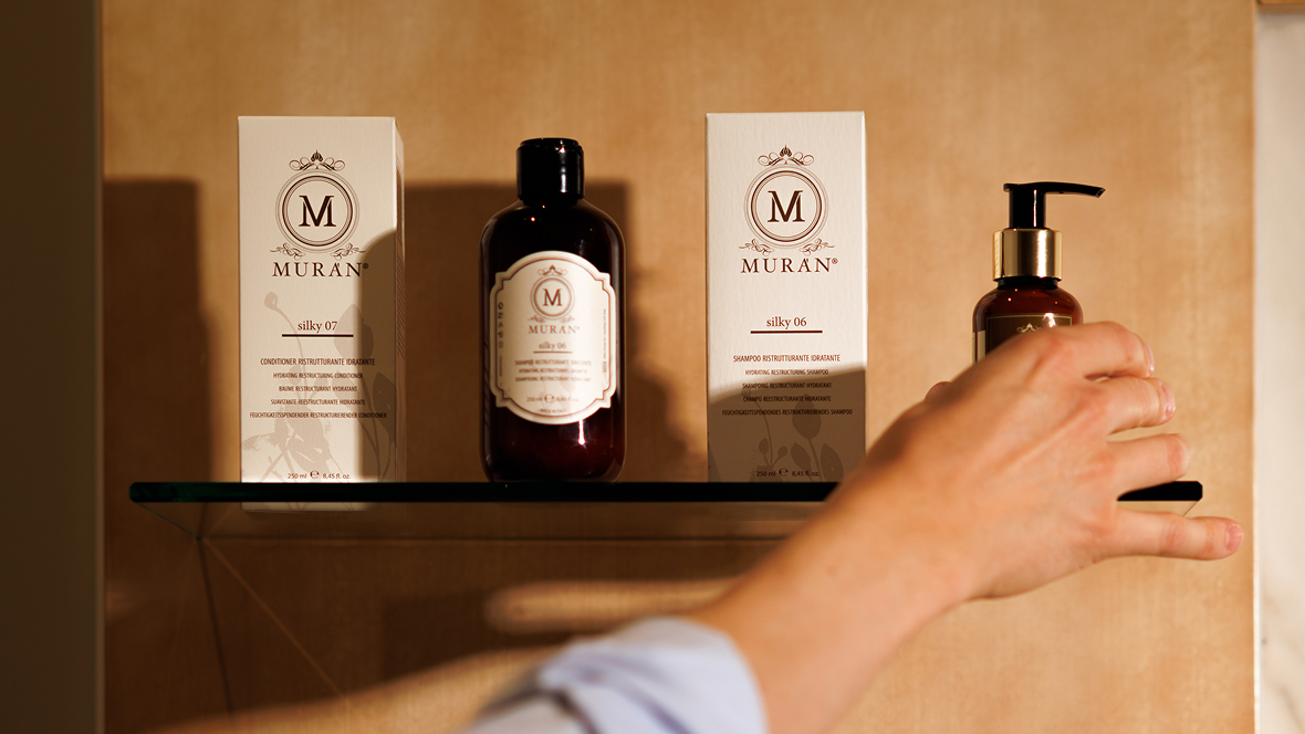 Hand reaching for a pump bottle of MURAN silky 06 shampoo on a glass shelf with MURAN product boxes and bottle under warm light.