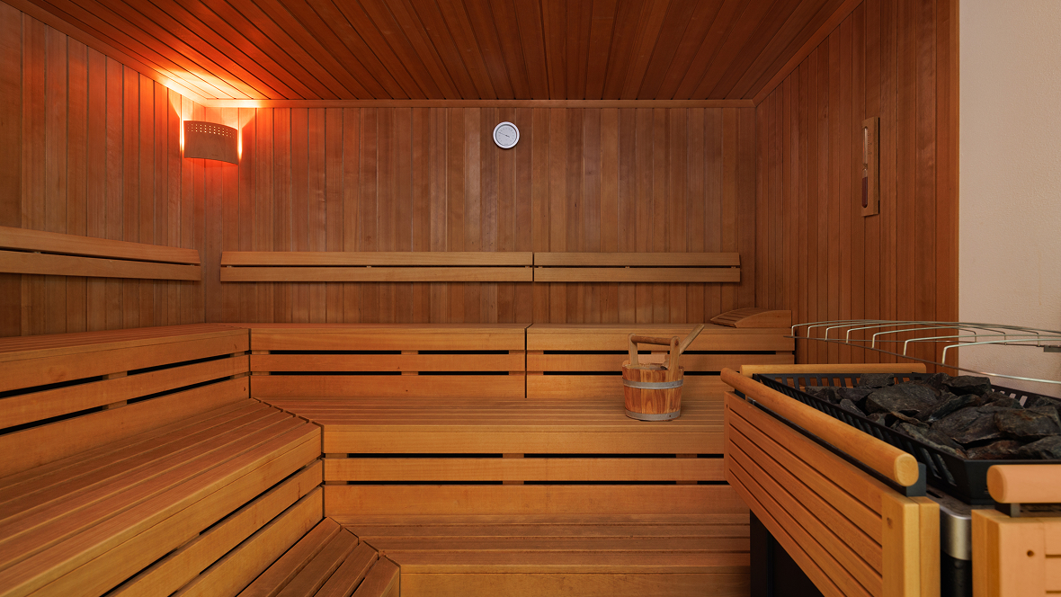 Innensauna mit Holzverkleidung, mehrstufigen Holzliegen, Eimer mit Kelle und Saunaofen mit Steinen.