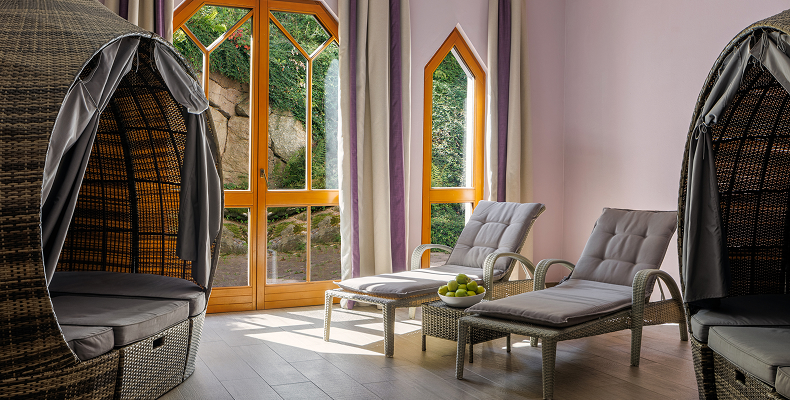 Gemütlicher Innenraum mit zwei Rattan-Loungestühlen mit grauen Polstern, einem kleinen Glasbeistelltisch und großen Fenstern mit Blick auf einen Garten.