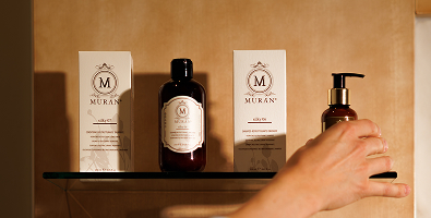 Zwei MURAN Hautpflegeprodukte und eine Hand, die eine Flasche mit Pumpspender auf einem Glasregal greift.