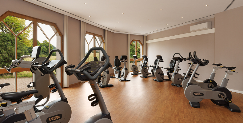 Innenraum eines Fitnessraums mit mehreren stationären Fahrrädern auf Holzboden und großen Fenstern mit Blick ins Grüne.