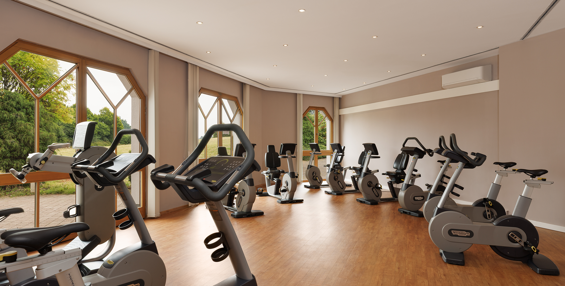 Fitnessraum mit mehreren stationären Fahrrädern vor großen Fenstern mit Blick auf Grünflächen.