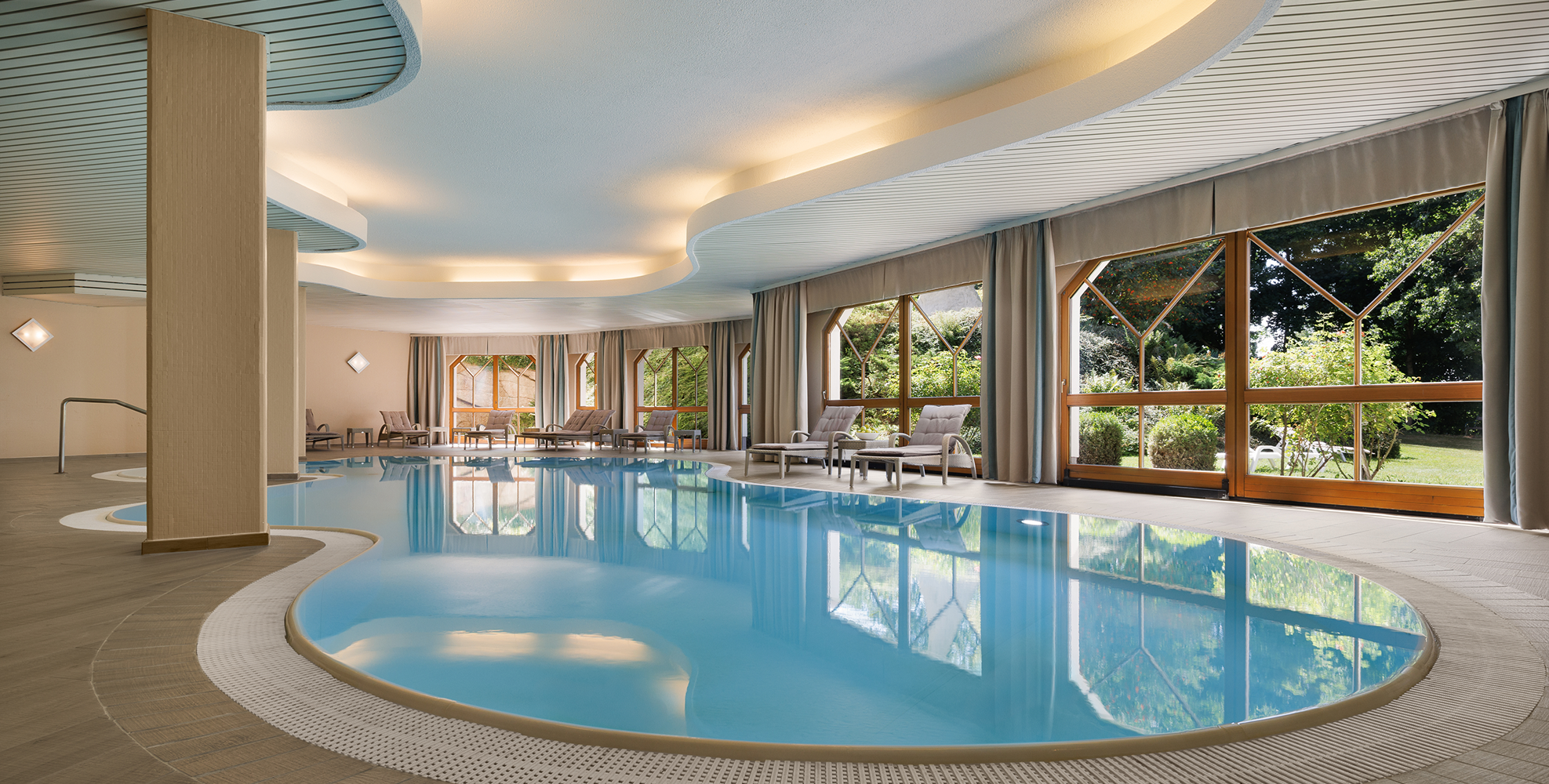 Innenaufnahme eines modernen Hallenbades mit blauer Wasserfläche, Komfortliegen am Rand und großen Fenstern mit Blick auf einen Garten.