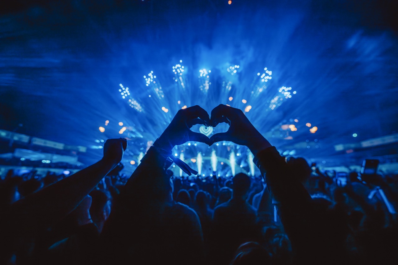 Silhouetted hands forming a heart shape against blue stage lights at a packed concert.