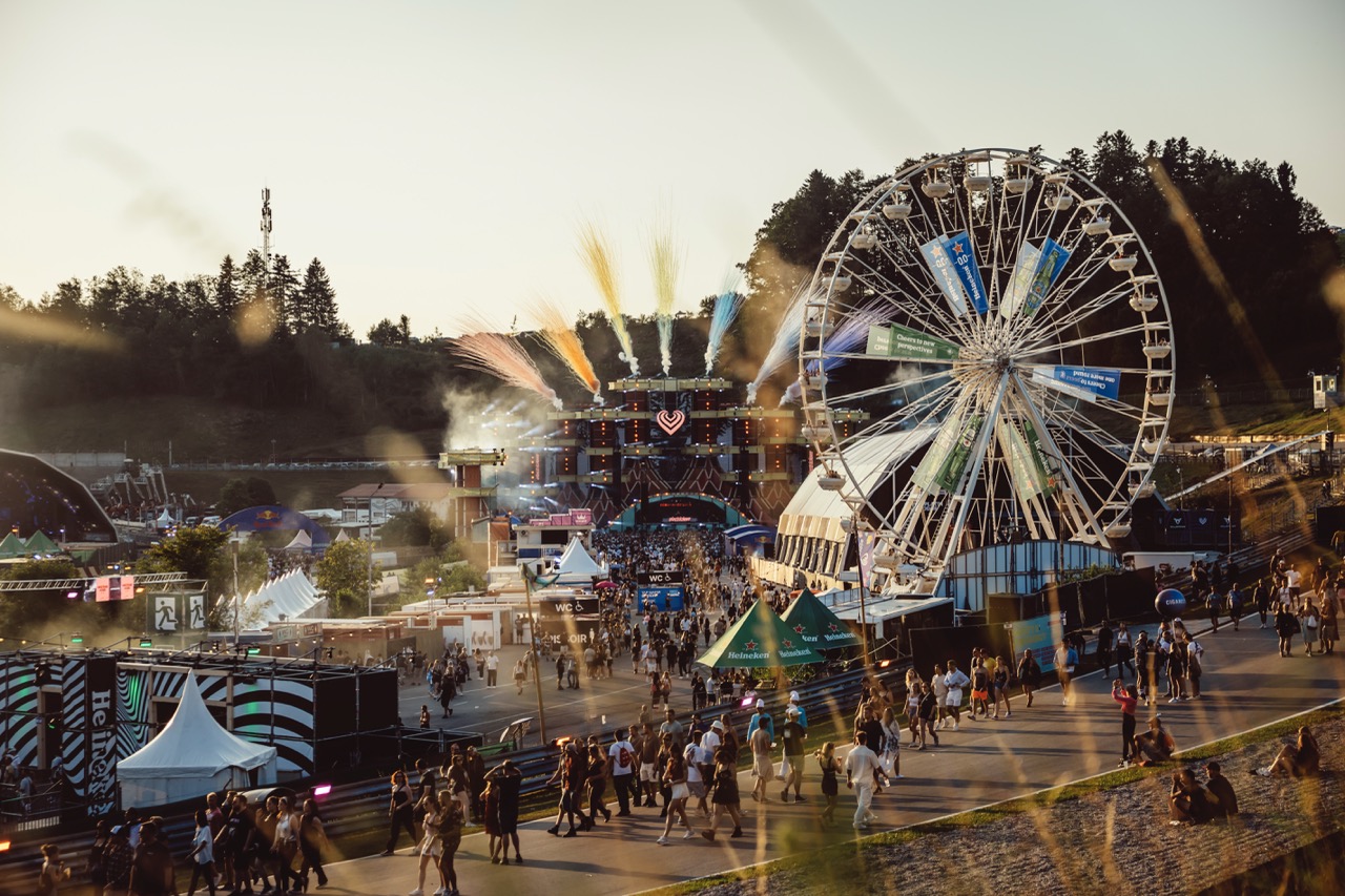 Crowd walking toward a large outdoor music festival stage with fireworks and a Ferris wheel during sunset.