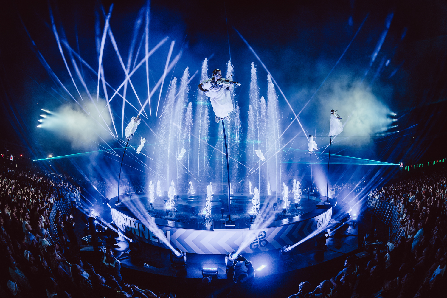Nighttime outdoor performance with musicians in white outfits suspended on poles above a fountain with water jets and blue stage lights, surrounded by a large audience.