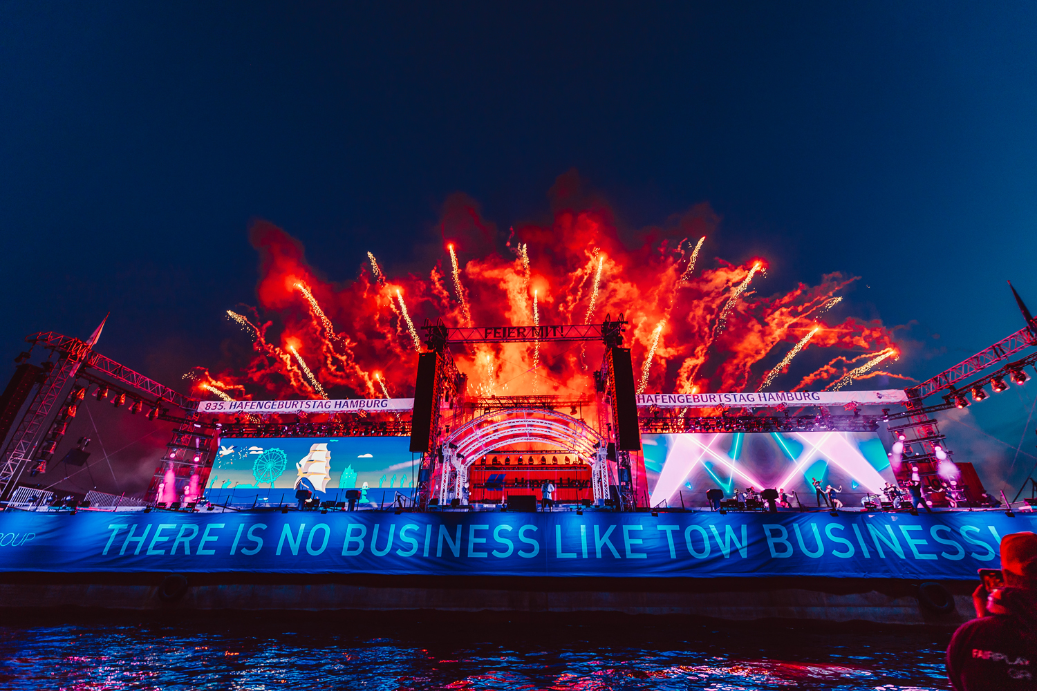Nighttime outdoor stage with fireworks bursting overhead and a banner reading 'There is no business like tow business!'