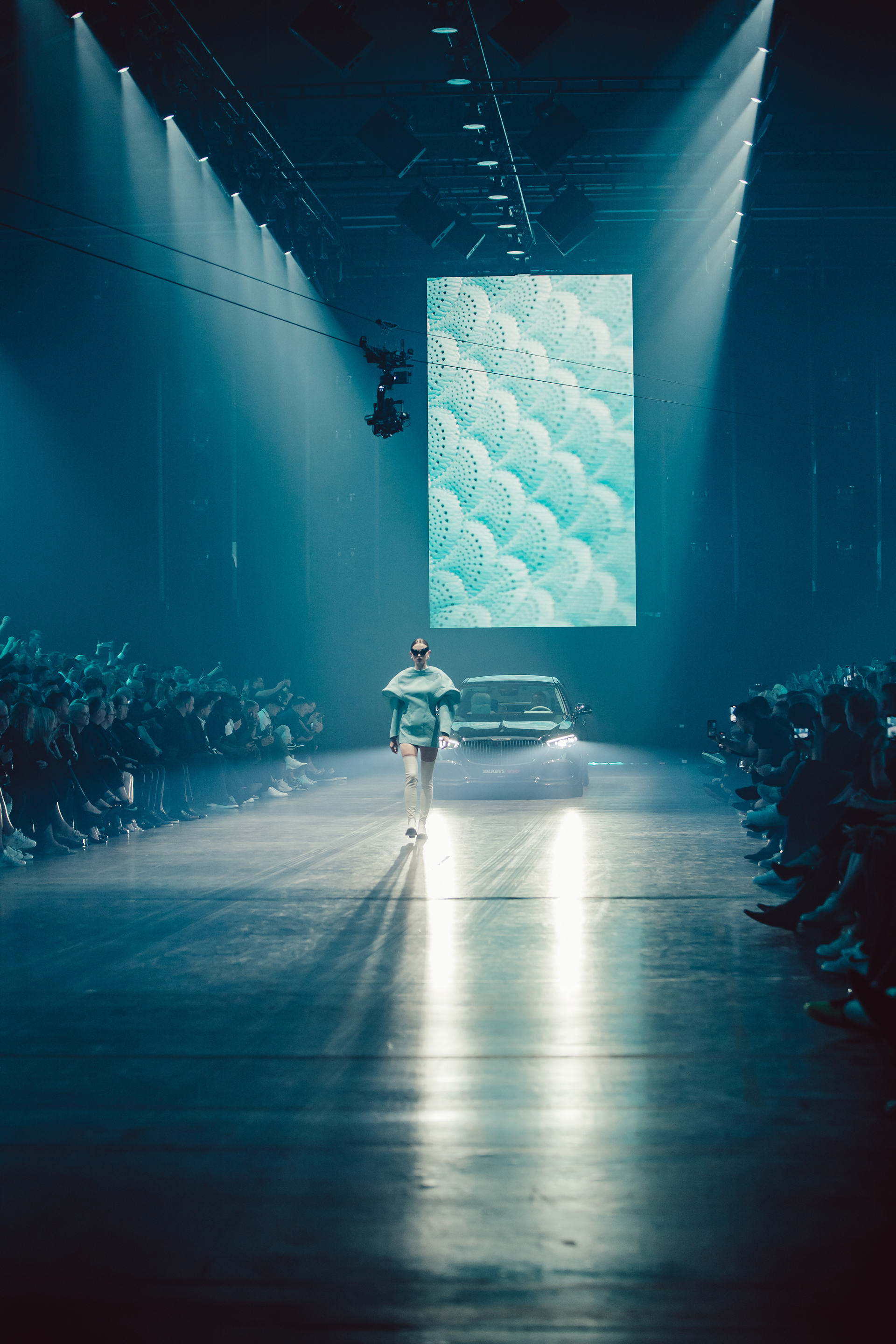 Fashion model wearing futuristic outfit walking on a runway with bright spotlights and a car behind under a large digital screen.