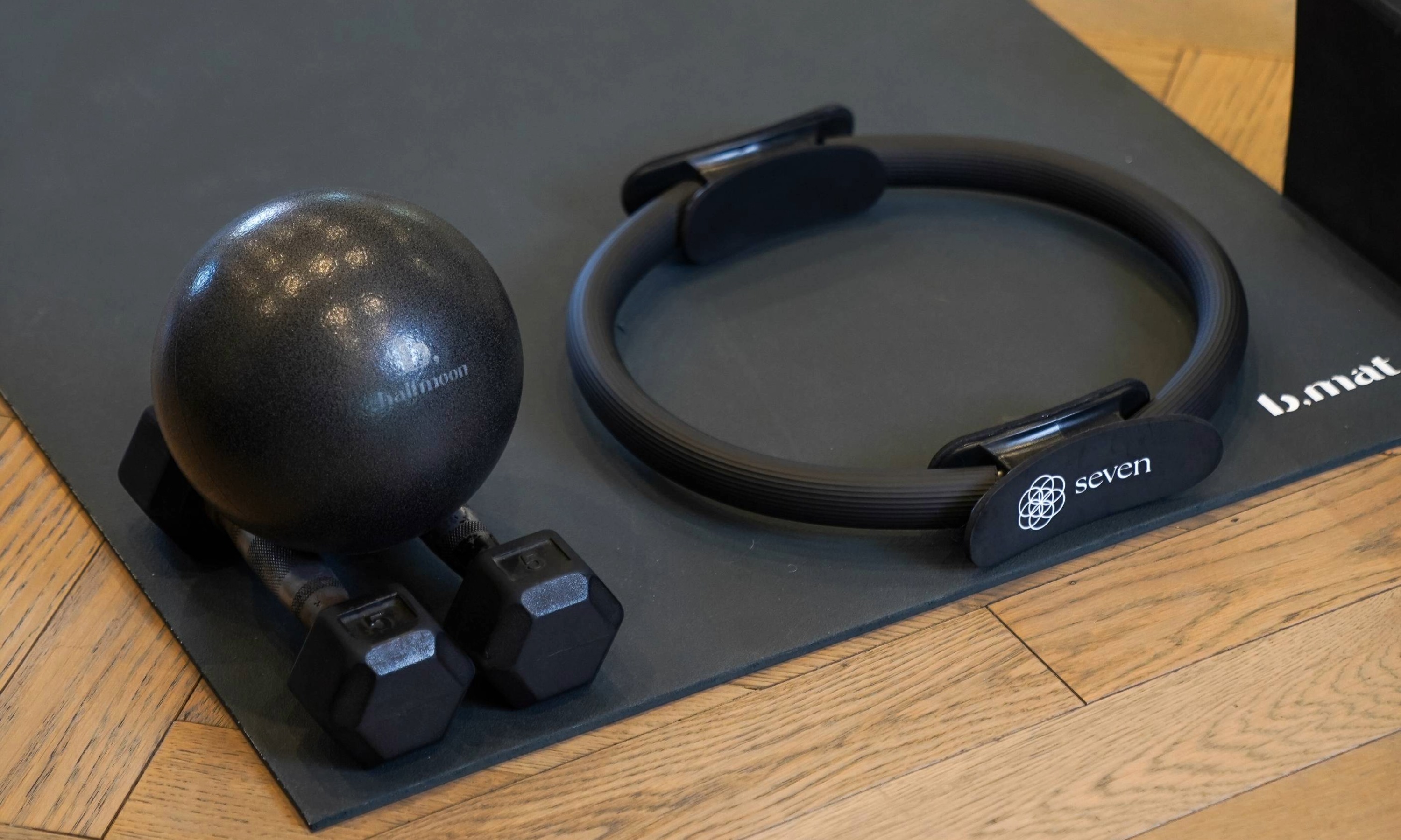 Black Pilates ring, small black Pilates ball, and two black dumbbells on a black yoga mat on wooden floor.