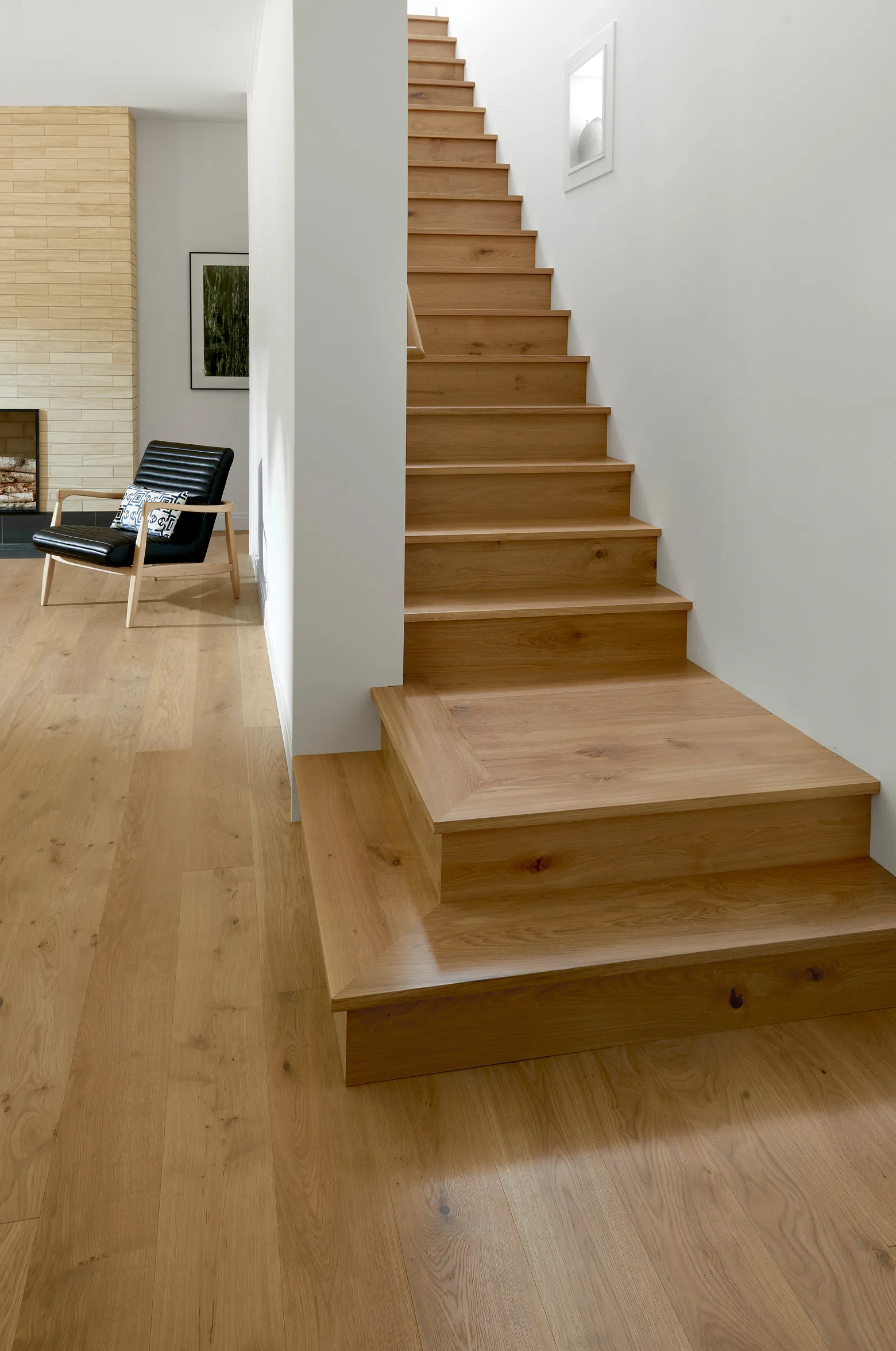 Wooden staircase with light wood flooring next to a black chair and a fireplace in a minimalist room.