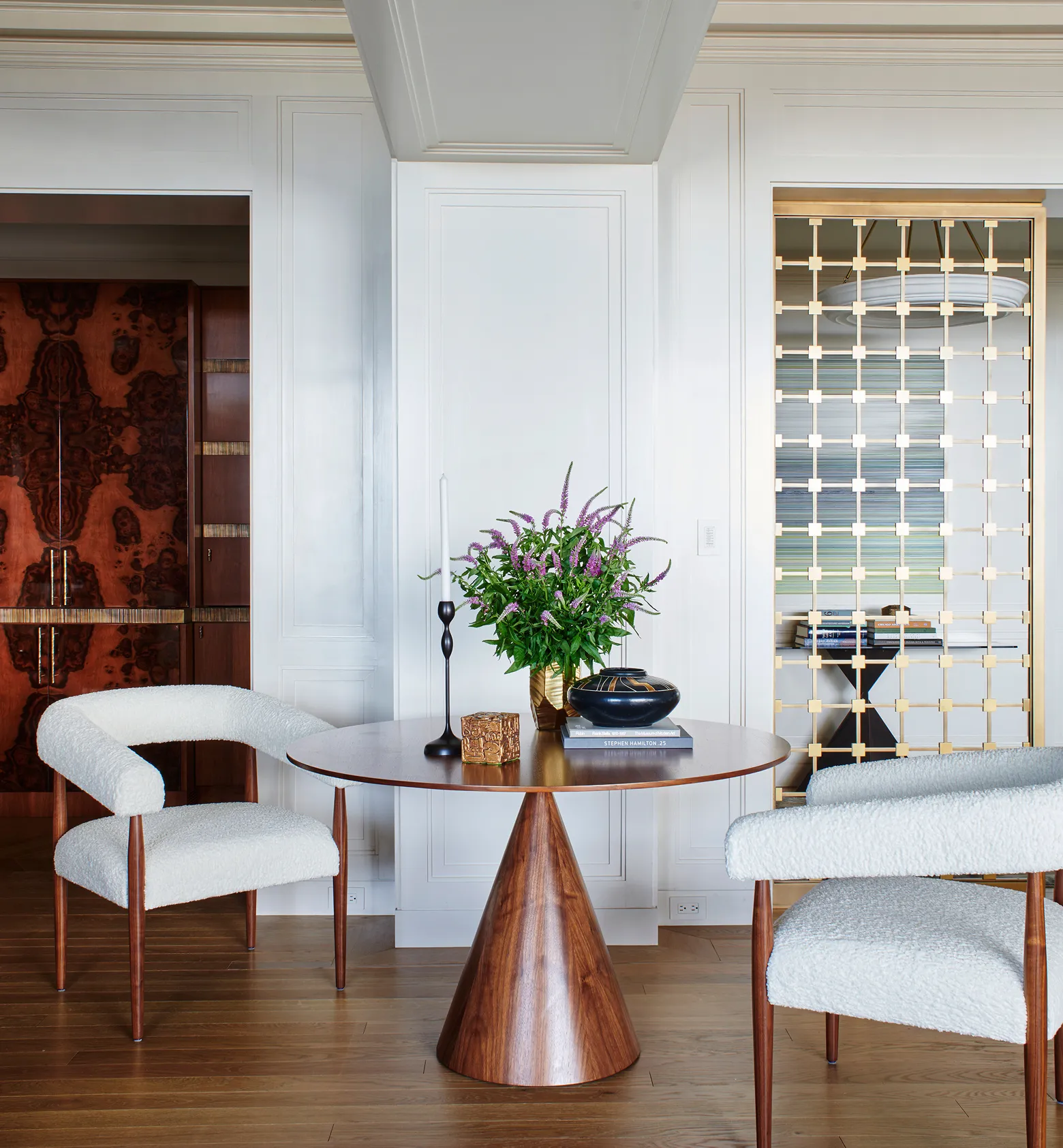 Round wooden table with flower arrangement, books, and two white upholstered chairs on wood floor.