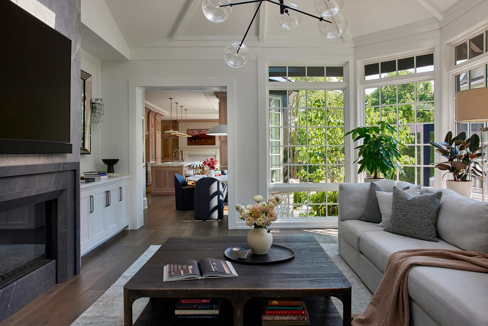 Bright living room with large windows, gray sofa, wooden coffee table, and view into kitchen dining area.