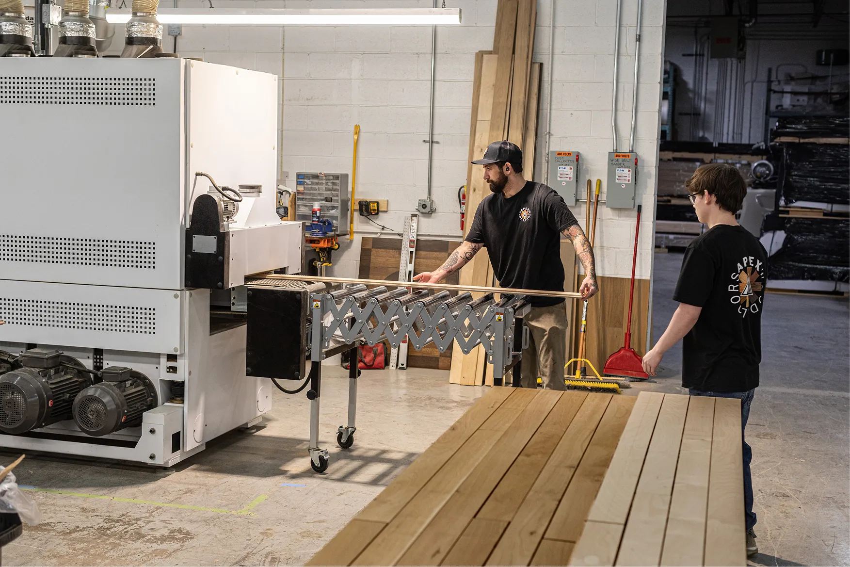 Two men working with wooden planks in a woodworking shop, operating a machine with rollers.