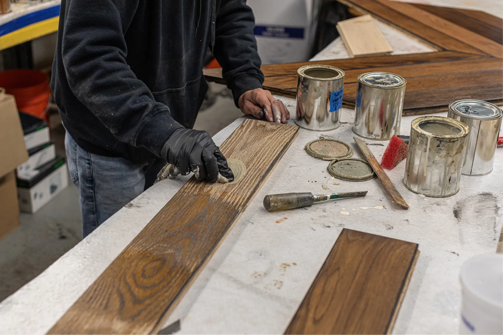 Person applying finish to wooden plank with cloth at a workbench with paint cans and tools.