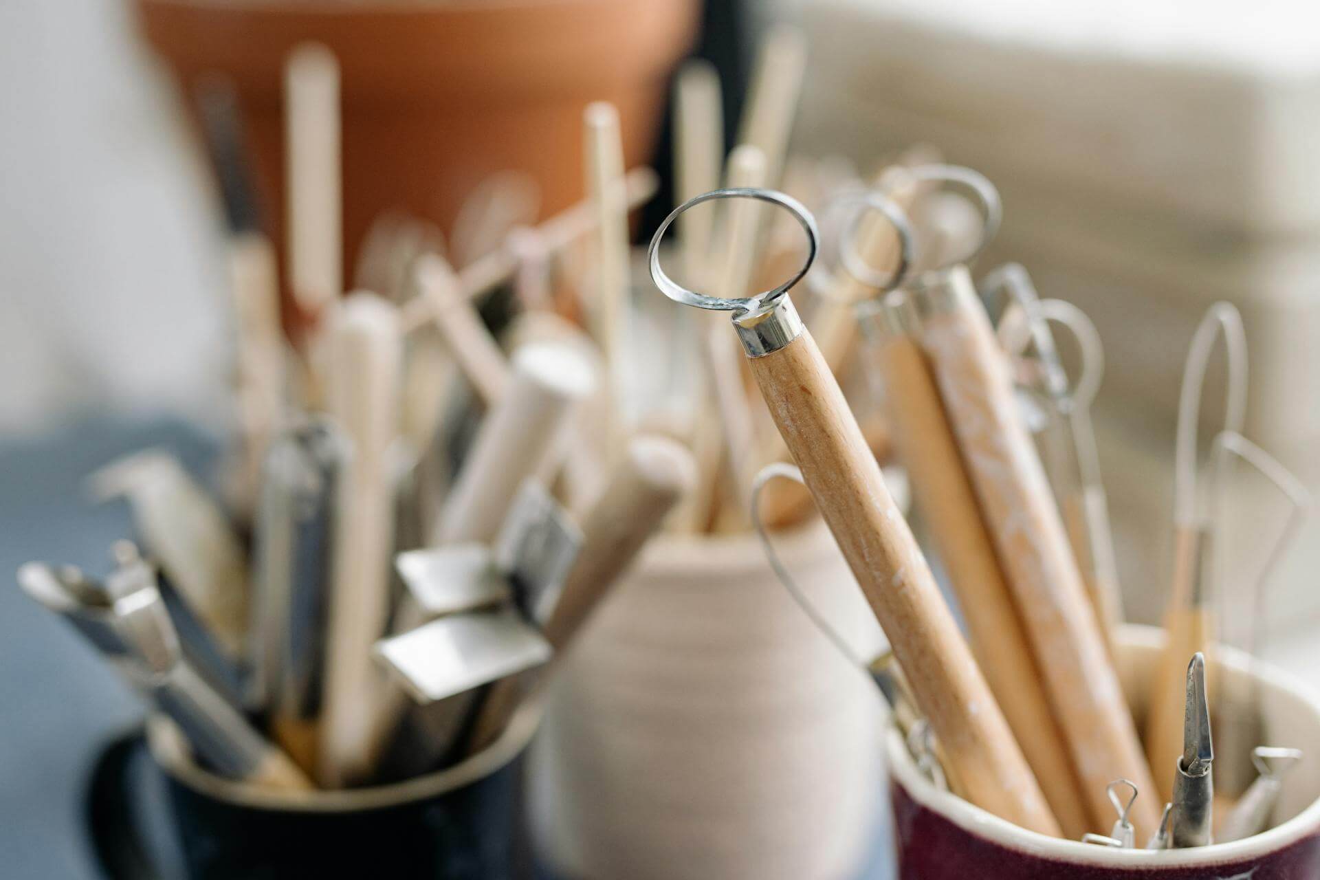 Close-up of pottery sculpting tools with wooden handles in cups on a work surface.