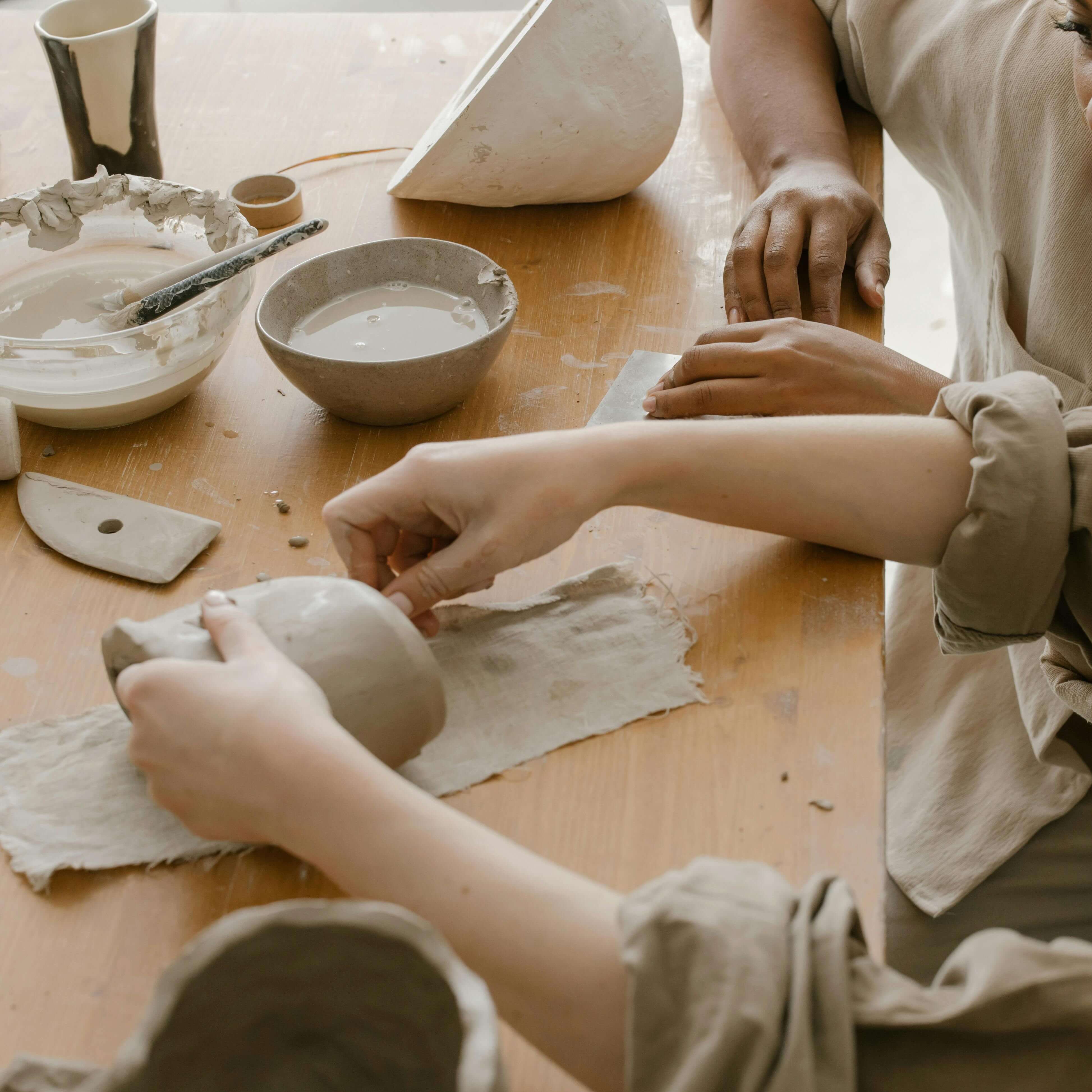 Two people working with clay on a wooden table, shaping pottery with their hands and tools.