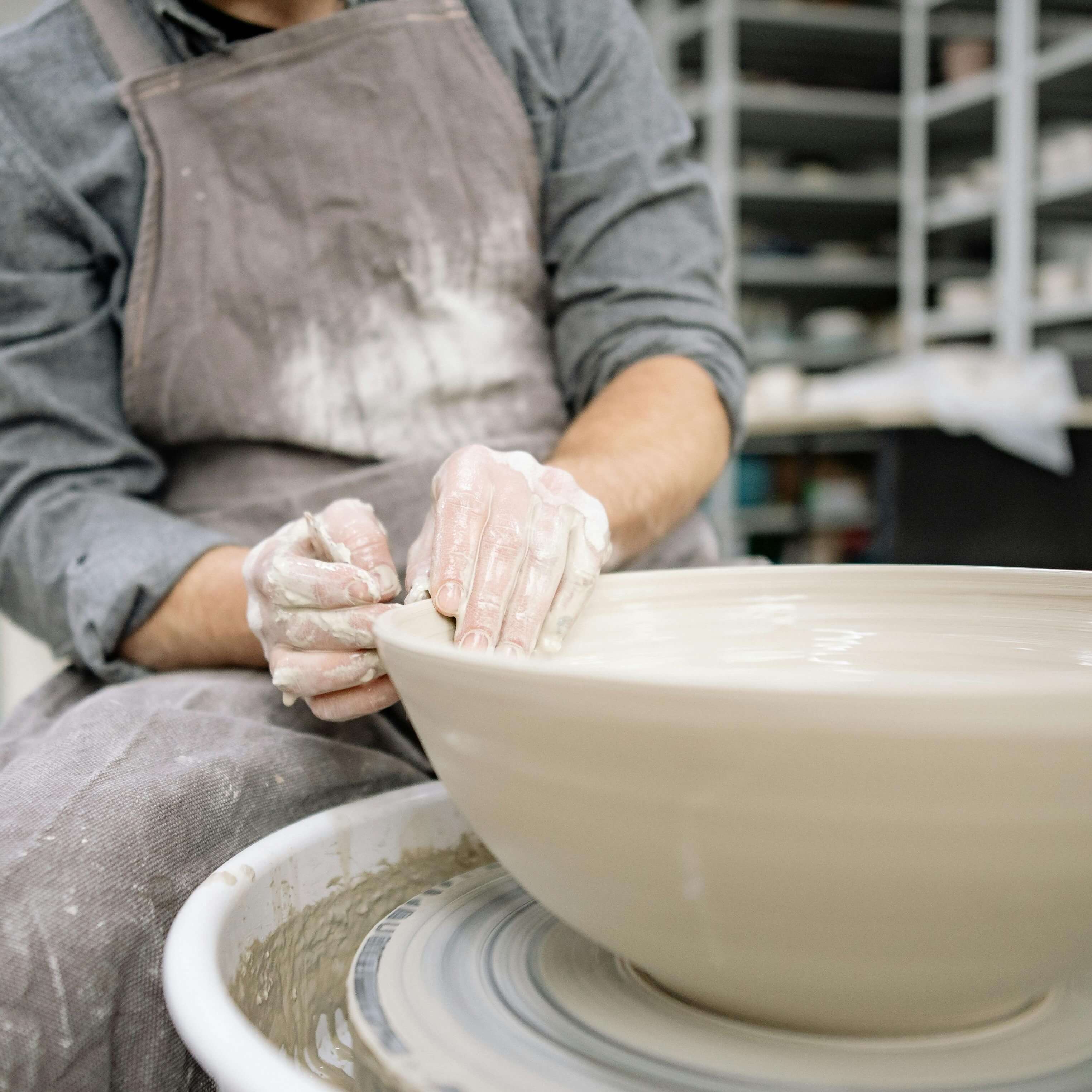 Potter shaping a large clay bowl on a spinning pottery wheel with hands covered in wet clay.