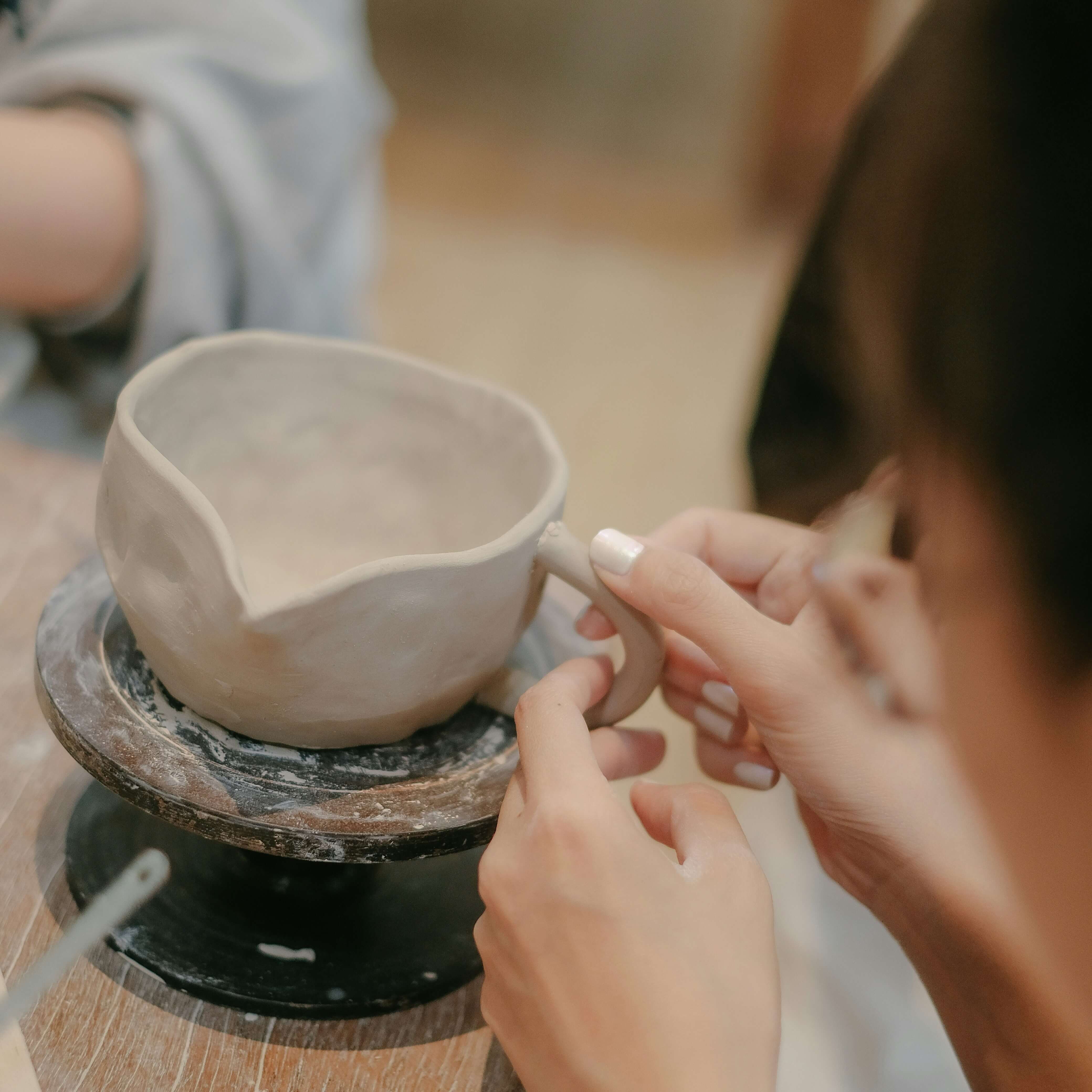 Hands shaping the handle of a handmade clay cup on a pottery wheel.