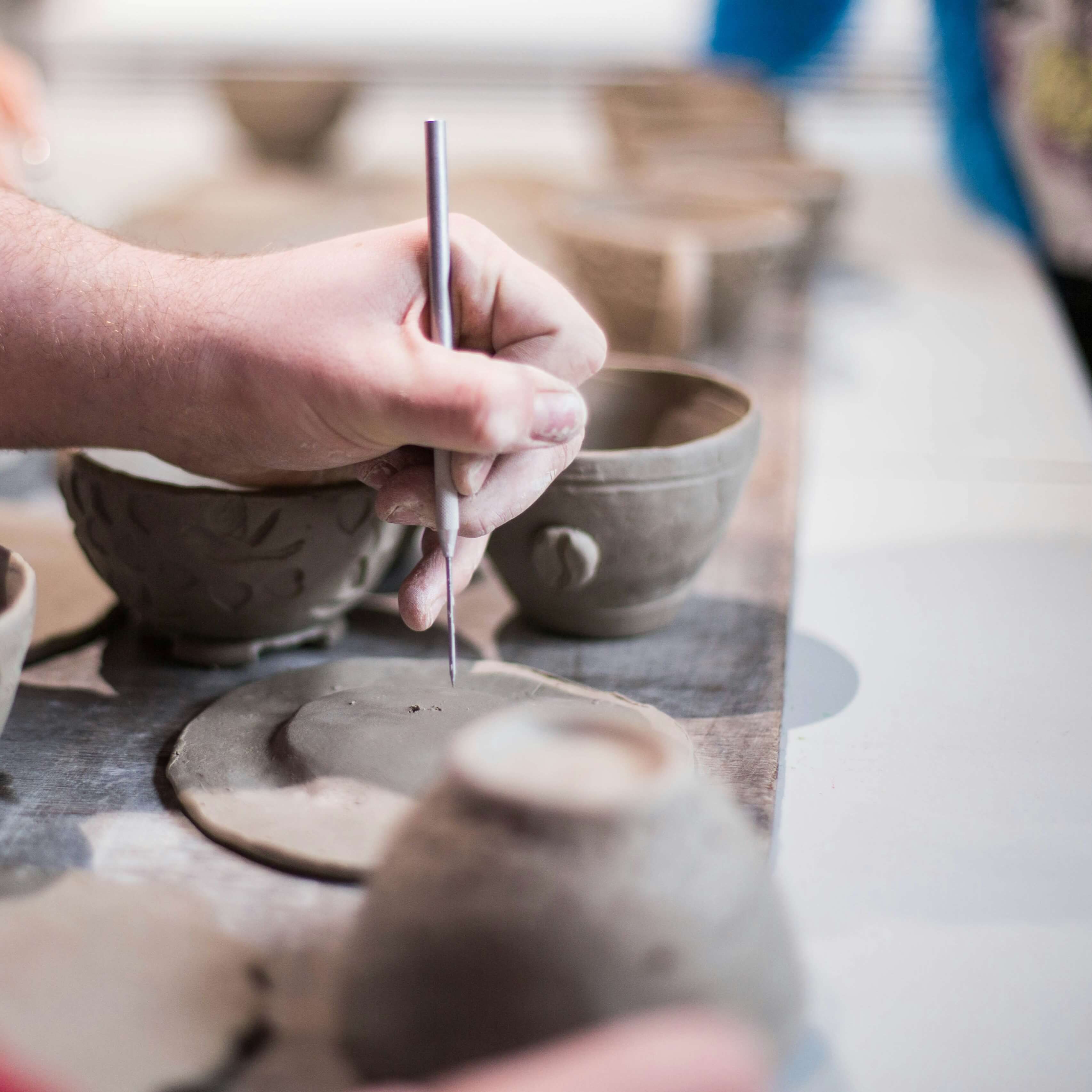 Close-up of a hand carving designs into clay pottery with a metal tool on a wooden surface.
