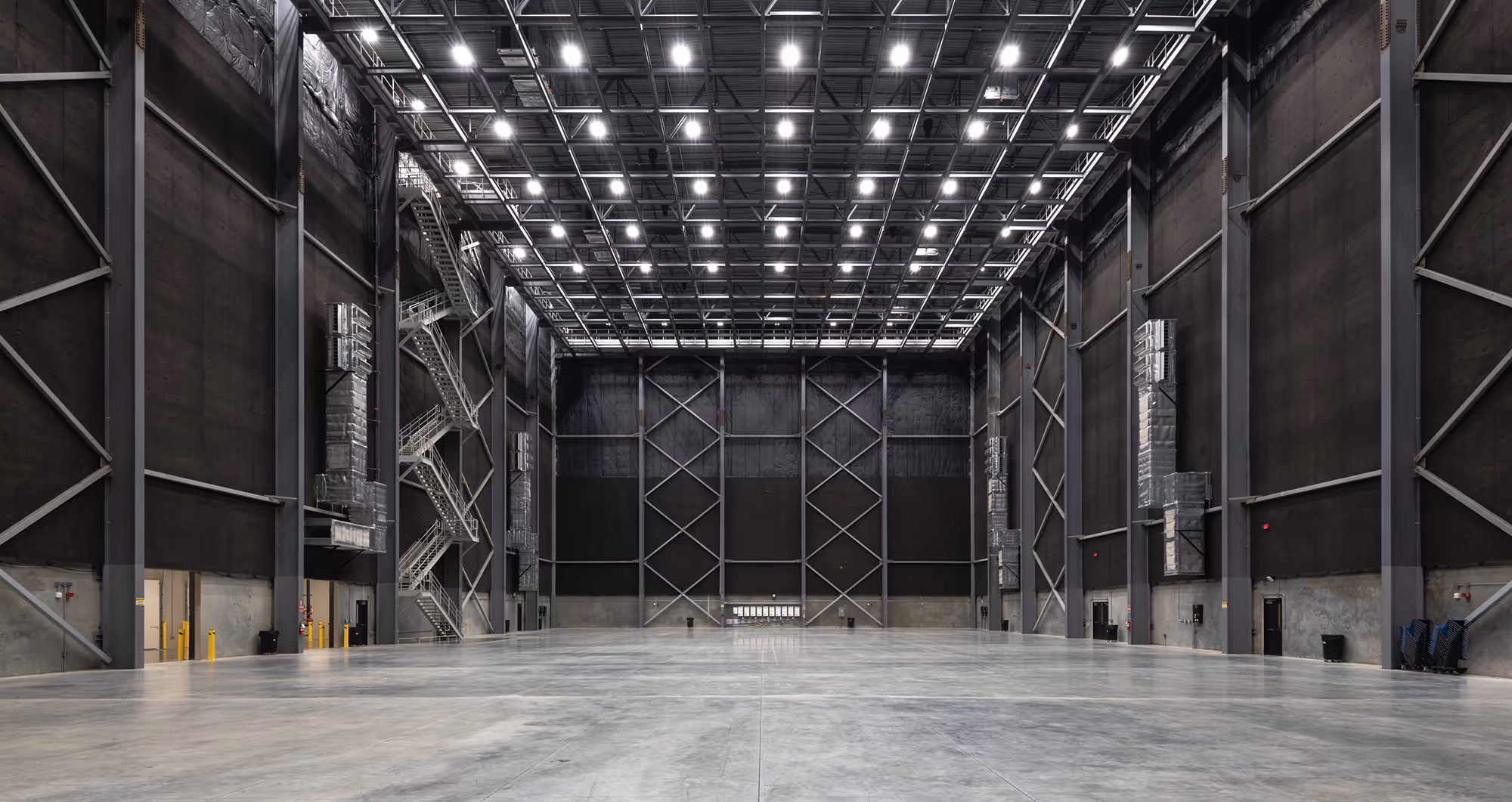 Large empty industrial rehearsal studio with high ceiling, metal beams, concrete floor, and bright overhead lights.