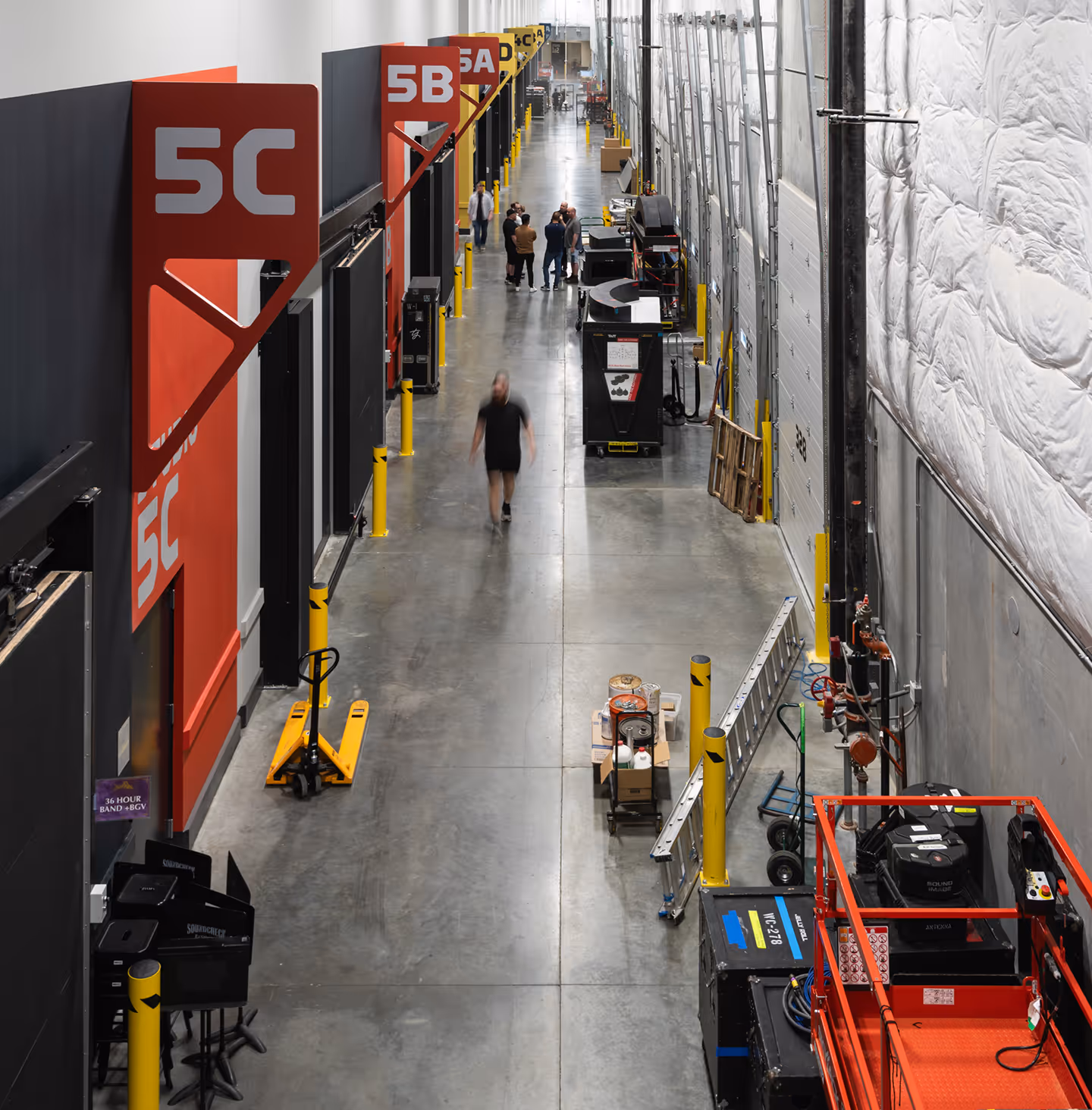 Long industrial corridor with numbered red and yellow room signs on left, equipment and materials on right, and people walking in the distance.