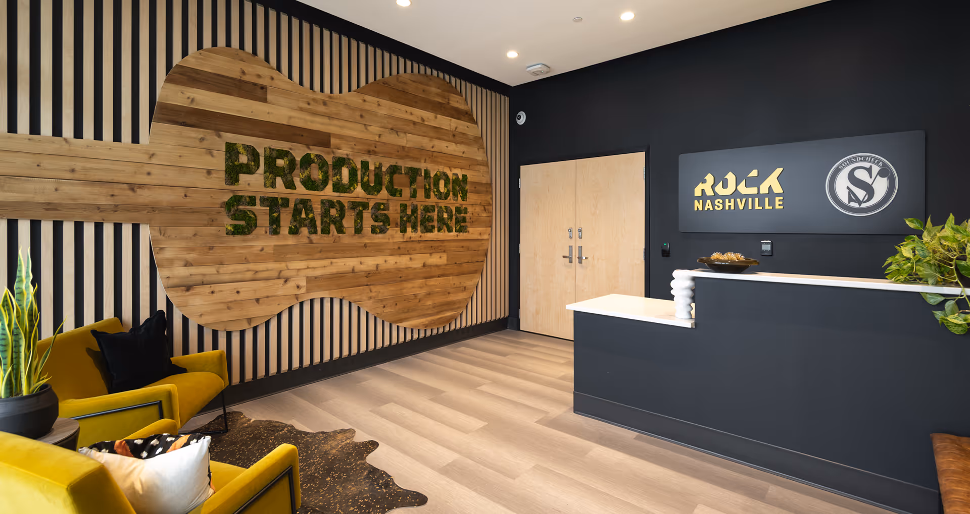 Modern reception area with dark walls, light wood flooring, yellow chairs, and a wooden wall sign reading 'PRODUCTION STARTS HERE'.