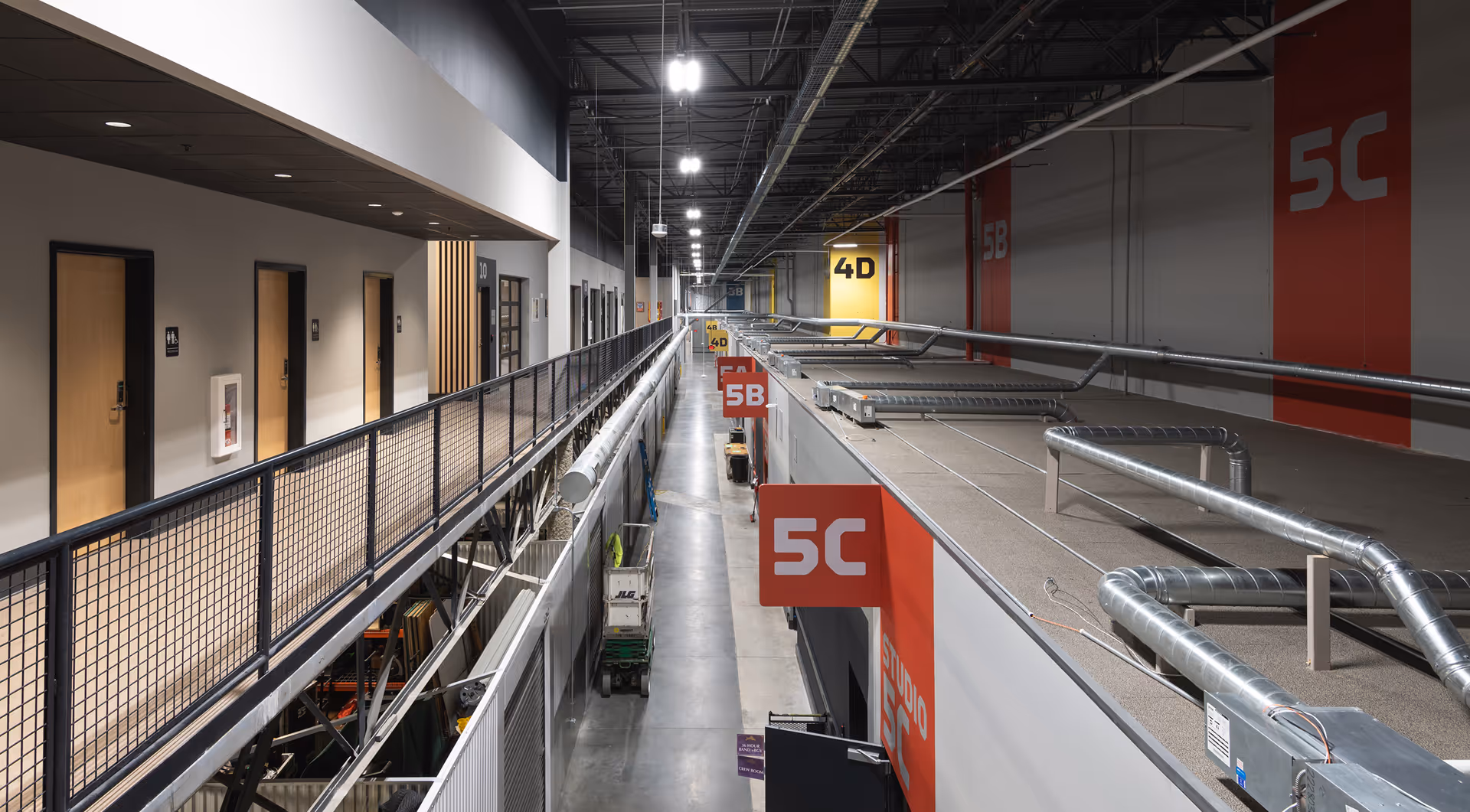 Interior view of a modern industrial hallway with numbered studio doors, metal railings, and exposed ducts overhead.