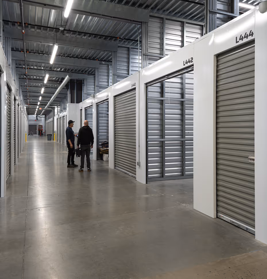 Two people standing in a brightly lit storage facility corridor with large metal roll-up doors numbered L440 to L444.