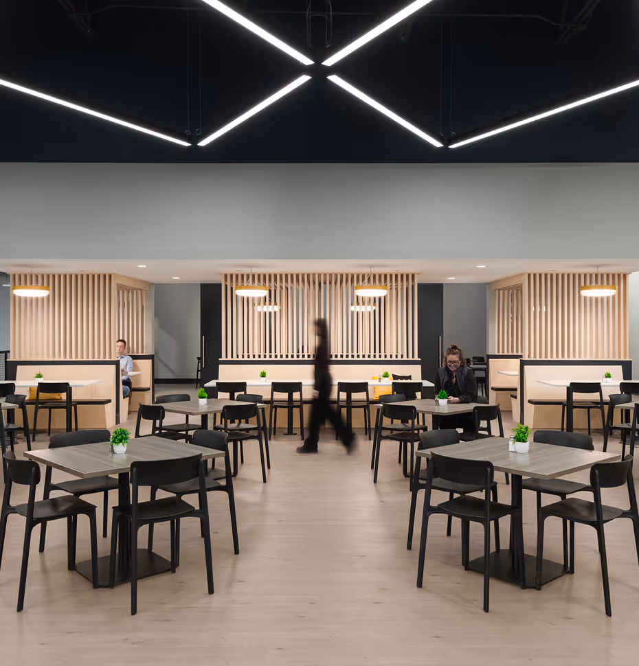 Modern cafeteria with wooden tables, black chairs, light wood-paneled booths, and geometric ceiling lights.